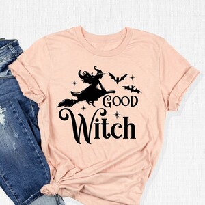 May include: A light pink t-shirt with the words "Good Witch" and a witch on a broomstick graphic. The shirt is paired with blue denim jeans. The t-shirt design includes bats and stars.
