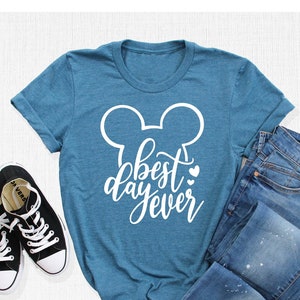 May include: A teal blue t-shirt with a white Mickey Mouse silhouette and the text "best day ever" in white cursive lettering. The shirt is laid flat on a white surface with a pair of black sneakers and a pair of blue jeans.