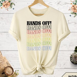 May include: A beige t-shirt with the text "HANDS OFF!" repeated in a rainbow of colors. The text is stacked vertically, with each line slightly offset from the one above it.