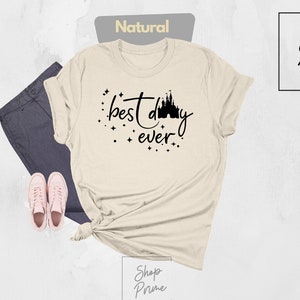 Puede incluir: Una camiseta beige con un gr&aacute;fico negro que dice "mejor d&iacute;a de todos" con una silueta del castillo de Cenicienta.