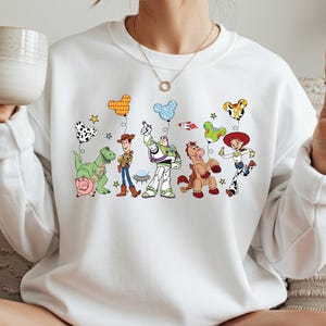 A woman is sitting on a couch, wearing a white sweatshirt with a graphic of various cartoon characters from the Toy Story franchise. She is holding a mug in her hand and making a peace sign with her fingers.