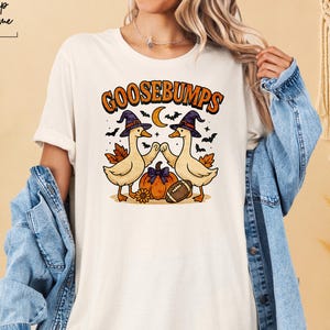 May include: Cream-colored t-shirt featuring a Halloween-themed graphic. The design includes two cartoon geese wearing witch hats, a pumpkin, a football, and the word "GOOSEBUMPS" in orange. The shirt also has the text "Shop Prime" in the upper left corner.