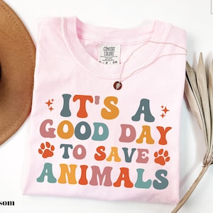 May include: A light pink t-shirt with the text "IT'S A GOOD DAY TO SAVE ANIMALS" in colorful, retro-style lettering. The shirt features paw prints and star graphics. A gold necklace with a pendant is draped on the shirt.