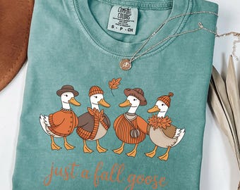 May include: Light green t-shirt with a crew neck and short sleeves. The shirt features a graphic of four cartoon ducks wearing fall attire, with the text "just a fall goose" in a cursive font. A silver necklace with a pendant is draped across the shirt.