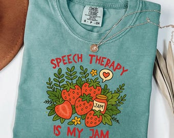 May include: Light green t-shirt with the text "SPEECH THERAPY IS MY JAM" in red, featuring a strawberry design. The shirt has a crew neck and short sleeves. A silver necklace with a pendant is draped across the shirt.