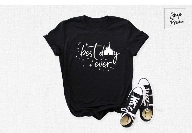 Puede incluir: Camiseta negra con texto blanco que dice "mejor d&iacute;a de todos" y una silueta blanca del castillo de Cenicienta.