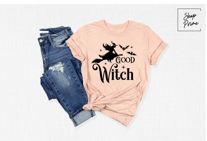 May include: A light pink t-shirt with the words "Good Witch" and a witch on a broomstick graphic. The shirt is paired with blue denim jeans. The t-shirt design includes bats and stars.