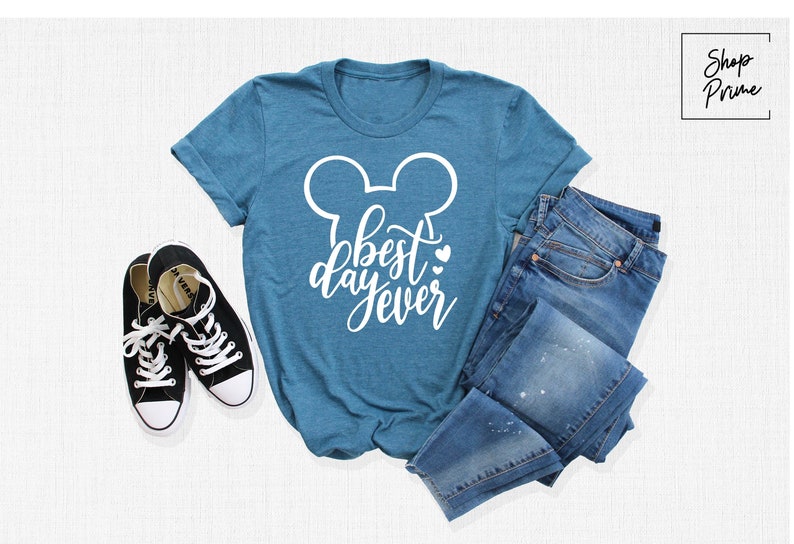 May include: A teal blue t-shirt with a white Mickey Mouse silhouette and the text "best day ever" in white cursive lettering. The shirt is laid flat on a white surface with a pair of black sneakers and a pair of blue jeans.