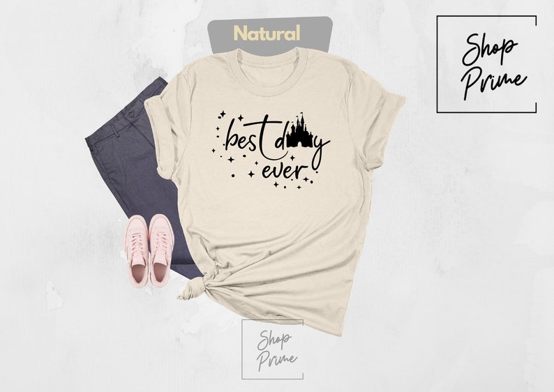 Puede incluir: Una camiseta beige con un gr&aacute;fico negro que dice "mejor d&iacute;a de todos" con una silueta del castillo de Cenicienta.