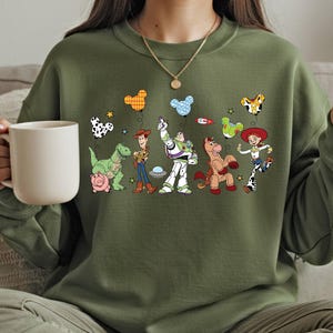 A woman wearing a green sweatshirt with a graphic of various cartoon characters from the Toy Story franchise, including Woody, Buzz Lightyear, and other beloved characters.