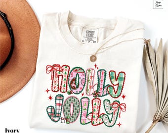 May include: Ivory-colored t-shirt with the words "HOLLY JOLLY" in a festive, patchwork design. The letters are decorated with Christmas patterns, bows, and stars. A gold necklace rests on the shirt.