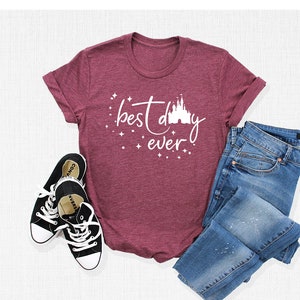 Puede incluir: Una camiseta de color granate con el texto "best day ever" y una silueta blanca del castillo de Disney. La camiseta est&aacute; sobre una superficie blanca con un par de zapatillas negras y unos vaqueros azules.