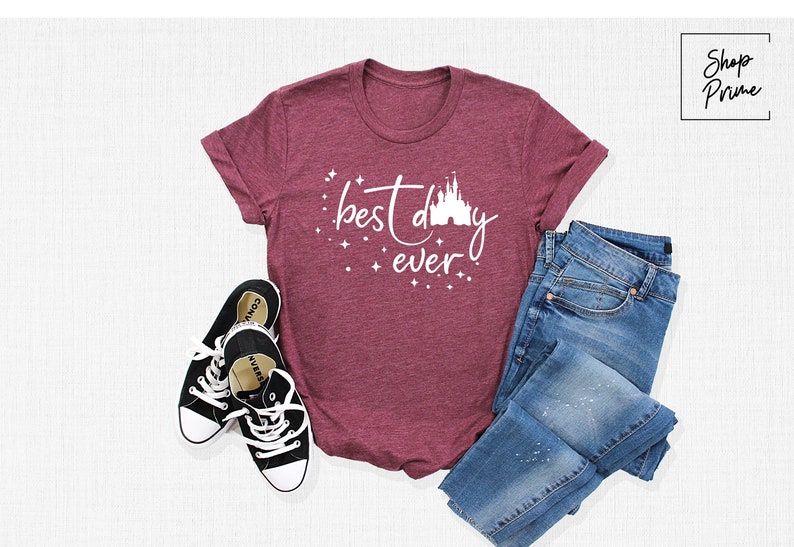 Puede incluir: Una camiseta de color granate con el texto "best day ever" y una silueta blanca del castillo de Disney. La camiseta est&aacute; sobre una superficie blanca con un par de zapatillas negras y unos vaqueros azules.