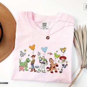 A pink t-shirt with a cartoon character design, including characters from the Toy Story franchise, such as Woody, Buzz Lightyear, and other beloved characters.