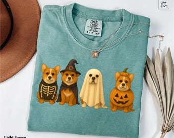 May include: Light green t-shirt featuring four cartoon corgi dogs dressed in Halloween costumes: a skeleton, a witch, a ghost, and a jack-o'-lantern. The shirt has a crew neck and short sleeves. A silver necklace is draped across the shirt.