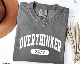 May include: A gray t-shirt with the text "OVERTHINKER 24/7" in white. The shirt is folded and has a silver necklace with a small pendant. A brown hat and dried plant are in the background.
