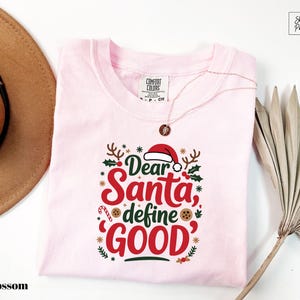 May include: A light pink t-shirt with the text "Dear Santa, define GOOD" in red and green. The design includes a Santa hat, holly, and gingerbread cookies. A gold necklace is draped over the shirt.