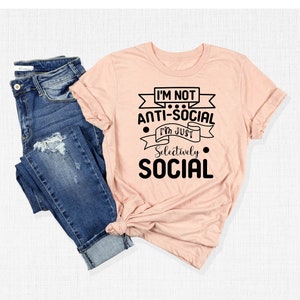May include: A peach-coloured t-shirt with black text that reads "I'm not anti-social I'm just selectively social". The shirt is folded and displayed with a pair of blue jeans.