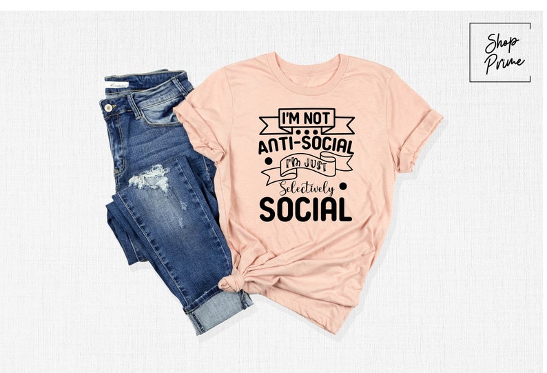 May include: A peach-coloured t-shirt with black text that reads "I'm not anti-social I'm just selectively social". The shirt is folded and displayed with a pair of blue jeans.