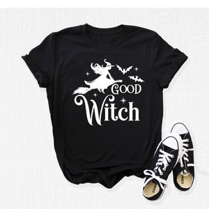 May include: Black t-shirt with a white graphic of a witch on a broom, bats, and the words "GOOD WITCH". Black and white sneakers are next to the shirt. The shirt is a Halloween-themed item.
