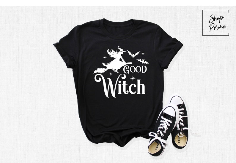 May include: Black t-shirt with a white graphic of a witch on a broom, bats, and the words "GOOD WITCH". Black and white sneakers are next to the shirt. The shirt is a Halloween-themed item.