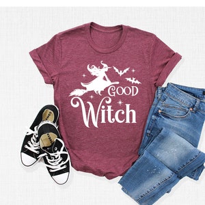 May include: Burgundy t-shirt with a white graphic of a witch on a broom, the words "Good Witch", and bats. The shirt is paired with blue jeans and black canvas sneakers. A Halloween-themed outfit.