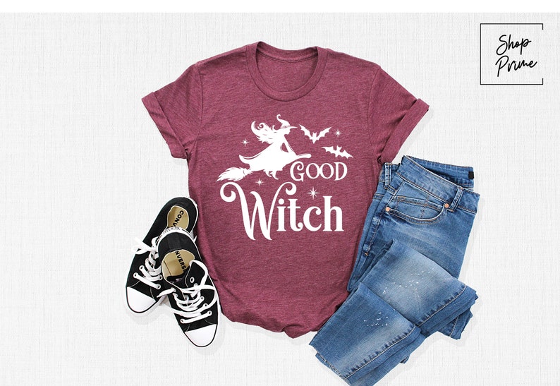 May include: Burgundy t-shirt with a white graphic of a witch on a broom, the words "Good Witch", and bats. The shirt is paired with blue jeans and black canvas sneakers. A Halloween-themed outfit.