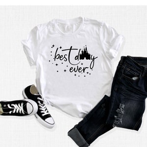 Puede incluir: Camiseta blanca con texto negro que dice "best day ever" y una silueta negra de un castillo. La camiseta est&aacute; sobre una superficie blanca con un par de zapatillas negras y unos vaqueros azules.