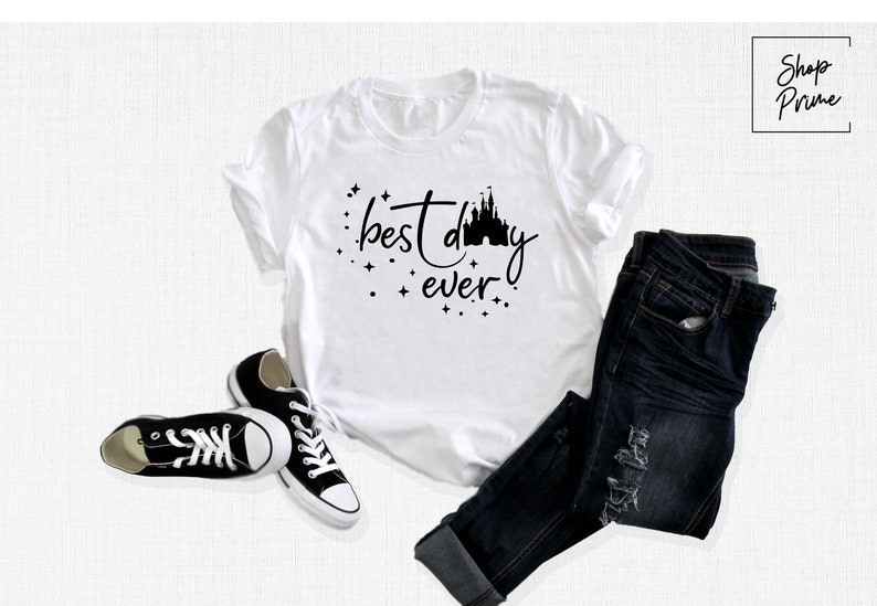 Puede incluir: Camiseta blanca con texto negro que dice "best day ever" y una silueta negra de un castillo. La camiseta est&aacute; sobre una superficie blanca con un par de zapatillas negras y unos vaqueros azules.