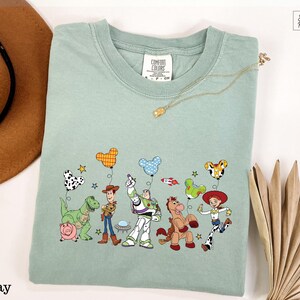 A green t-shirt with a graphic of characters from the Toy Story franchise, including Woody, Buzz Lightyear, and other beloved characters, along with various Disney-themed elements such as balloons and stars.