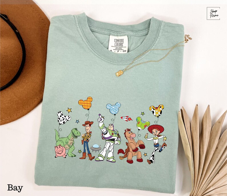 A green t-shirt with a graphic of characters from the Toy Story franchise, including Woody, Buzz Lightyear, and other beloved characters, along with various Disney-themed elements such as balloons and stars.