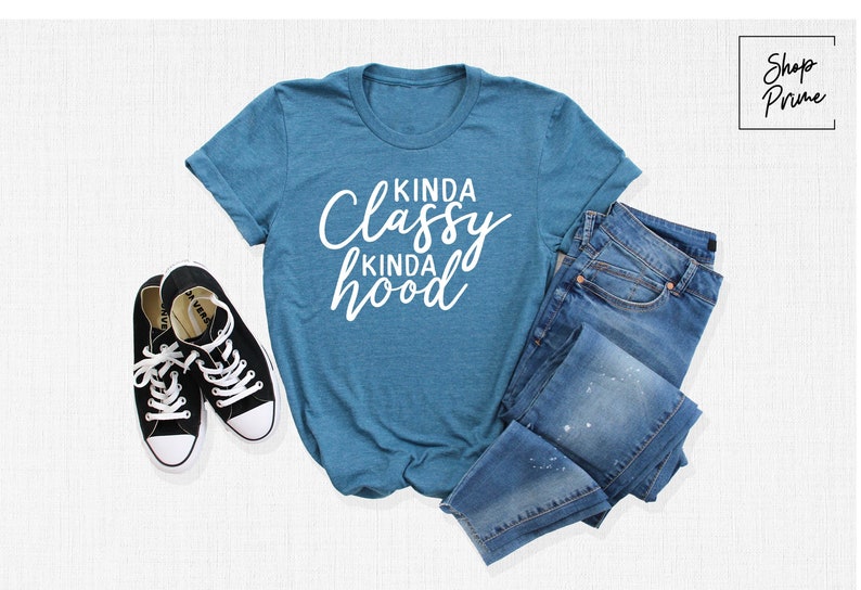 May include: A teal blue t-shirt with the text "Kinda Classy Kinda Hood" in white lettering. The shirt is laid flat on a white surface with a pair of black sneakers and a pair of blue jeans.