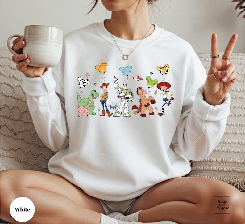 A woman is sitting on a couch, wearing a white sweatshirt with a graphic of various cartoon characters from the Toy Story franchise. She is holding a mug in her hand and making a peace sign with her fingers.