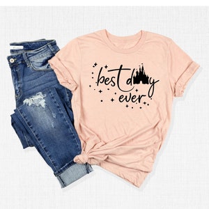 Puede incluir: Una camiseta rosa con un gr&aacute;fico negro que dice "mejor d&iacute;a de todos" con una silueta de un castillo.