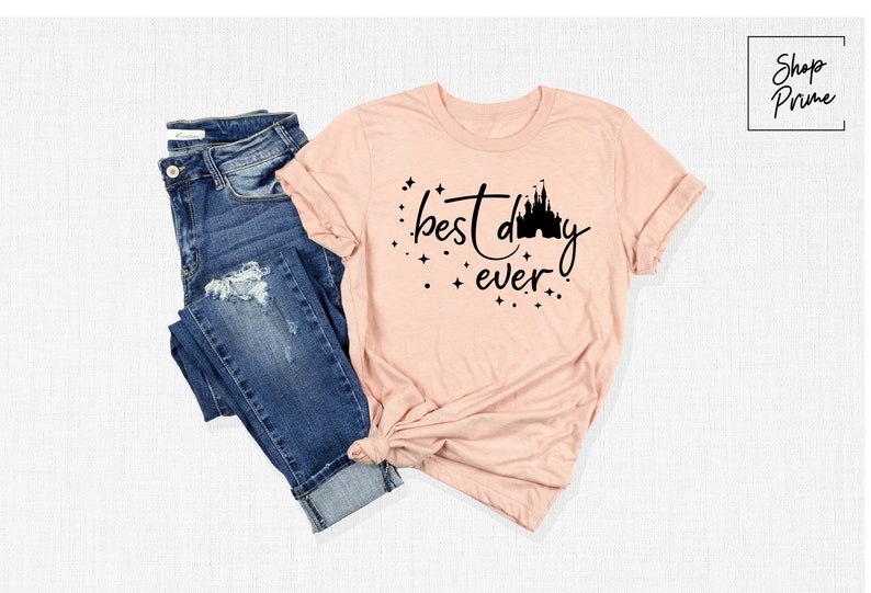 Puede incluir: Una camiseta rosa con un gr&aacute;fico negro que dice "mejor d&iacute;a de todos" con una silueta de un castillo.