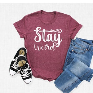 以下が含まれることがあります： 白い文字で「Stay Weird」と書かれたマルーンのTシャツ。Tシャツは白い表面に黒のスニーカーと青いジーンズと一緒に平らに置かれています。