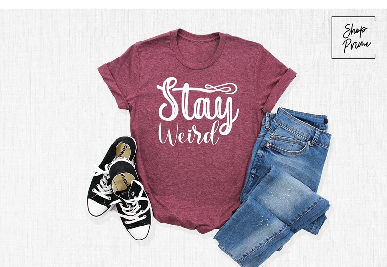 以下が含まれることがあります： 白い文字で「Stay Weird」と書かれたマルーンのTシャツ。Tシャツは白い表面に黒のスニーカーと青いジーンズと一緒に平らに置かれています。