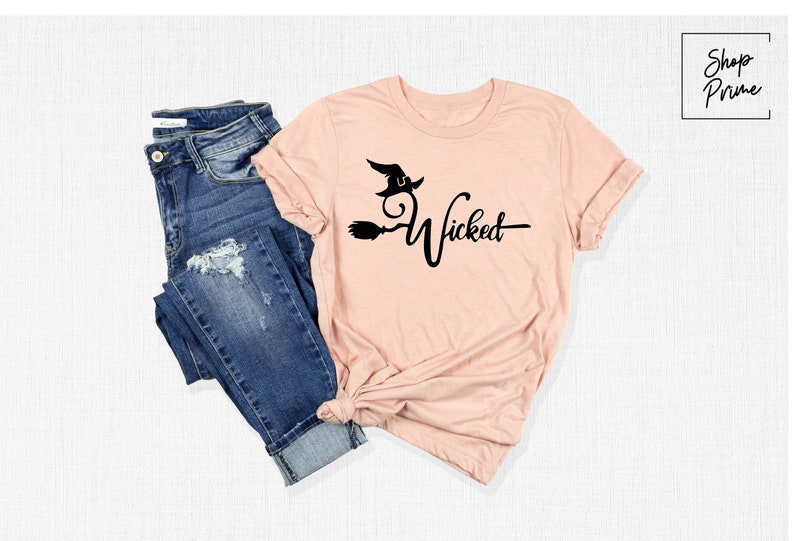 Puede incluir: Una camiseta rosa con una silueta de bruja negra y la palabra "Wicked" impresa en ella. La camiseta est&aacute; doblada y se muestra con un par de pantalones vaqueros azules.