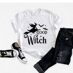 May include: White t-shirt with a black graphic of a witch on a broomstick, bats, and the words "Good Witch." The shirt is paired with dark blue jeans and black canvas sneakers. A Halloween-themed outfit.