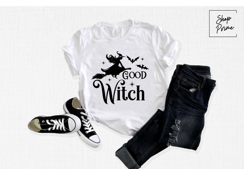 May include: White t-shirt with a black graphic of a witch on a broomstick, bats, and the words "Good Witch." The shirt is paired with dark blue jeans and black canvas sneakers. A Halloween-themed outfit.