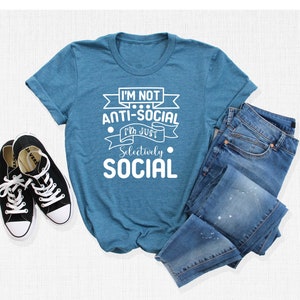 May include: A teal blue t-shirt with white text that reads "I'm not anti-social I'm just selectively social". The shirt is laid flat on a white surface with a pair of black trainers and a pair of blue jeans.