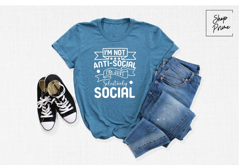 May include: A teal blue t-shirt with white text that reads "I'm not anti-social I'm just selectively social". The shirt is laid flat on a white surface with a pair of black trainers and a pair of blue jeans.