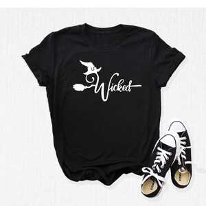 Puede incluir: Camiseta negra con un gr&aacute;fico blanco de una bruja volando en una escoba con el texto "Wicked".