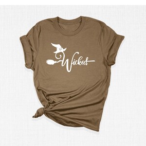 Puede incluir: Una camiseta marr&oacute;n con un gr&aacute;fico blanco de una bruja volando en una escoba con el texto "Wicked".