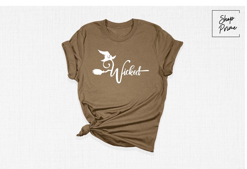 Puede incluir: Una camiseta marr&oacute;n con un gr&aacute;fico blanco de una bruja volando en una escoba con el texto "Wicked".