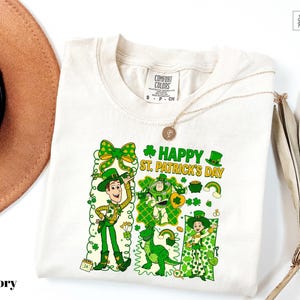 A white t-shirt with a colorful graphic design depicting cartoon characters and the text "Happy St. Patrick's Day" printed on it.