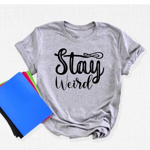 以下が含まれることがあります： 前面に黒い文字で「Stay Weird」とプリントされた灰色のTシャツです。
