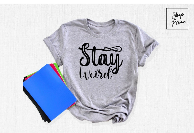 以下が含まれることがあります： 前面に黒い文字で「Stay Weird」とプリントされた灰色のTシャツです。