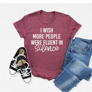 Pu&ograve; includere: Una maglietta color granata con il testo "I wish more people were fluent in silence" in lettere bianche. La maglietta &egrave; stesa su una superficie bianca con un paio di scarpe da ginnastica nere e un paio di jeans blu.