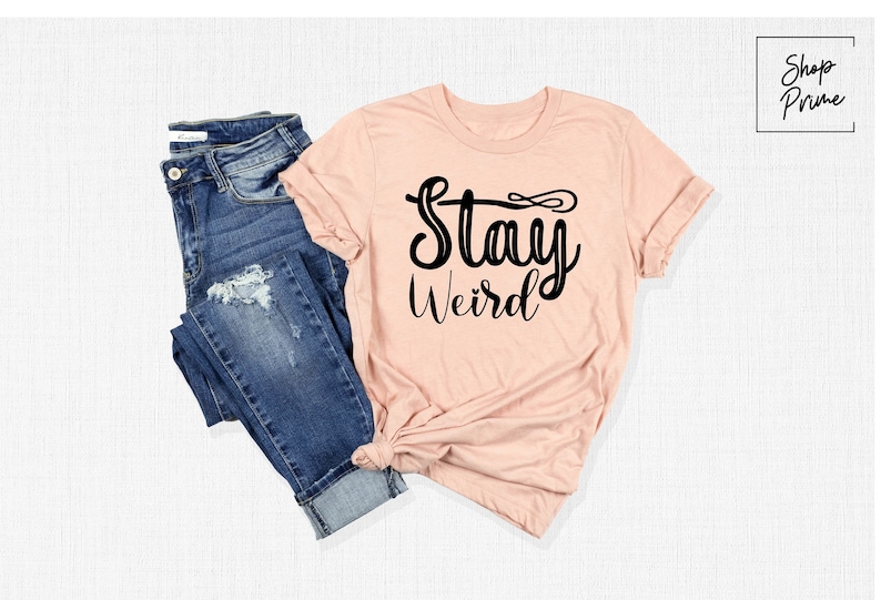 以下が含まれることがあります： 白地に桃色のTシャツに、黒い筆記体で「Stay Weird」と書いてあります。Tシャツは白い表面に置かれ、青いジーンズが一緒に置かれています。